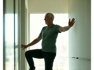 Starszy mężczyzna utrzymuje równowagę na jednej nodze, wspierając się ręką na ścianie, symbolizując ćwiczenia równowagi.